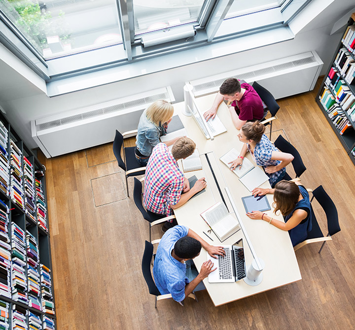 Group of people working at a shared table on laptops with bookshelves.